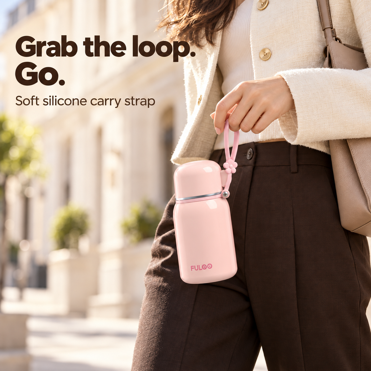 An adult carrying a Blush Pink FULOO travel bottle by its soft silicone loop on a sunlit city street.